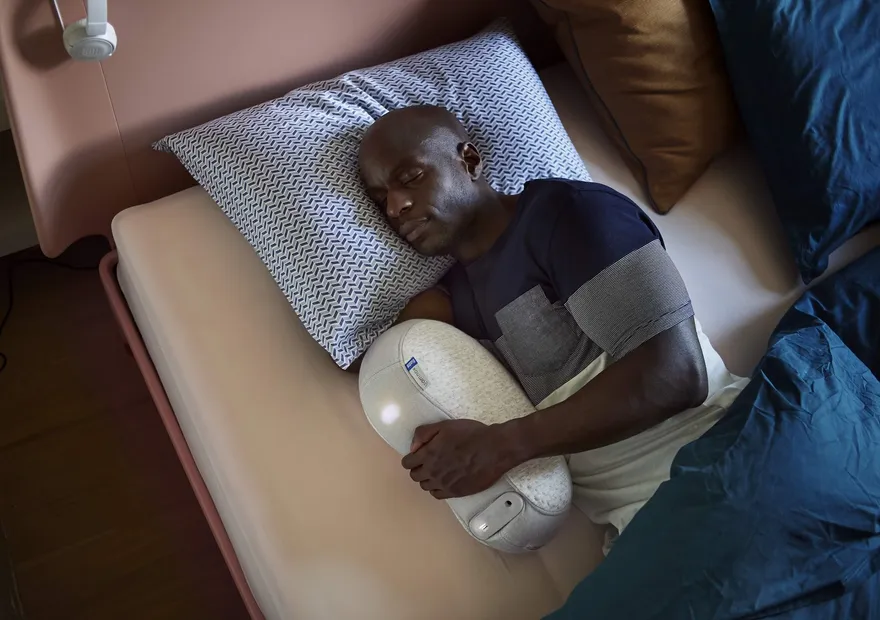 A man asleep on a bed hugging a white smart pillow, seen from above