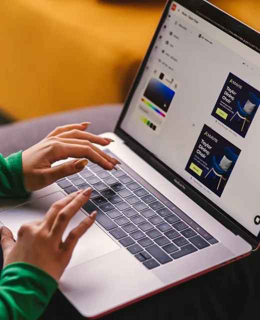 Hands typing on a laptop that shows ad layouts for a dining chair