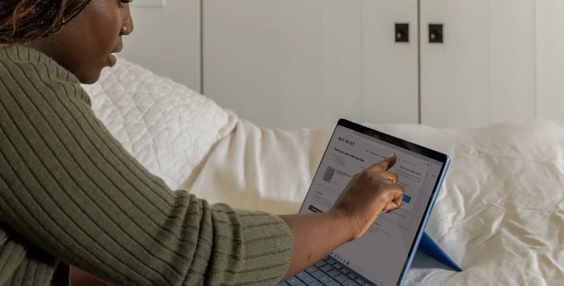 A woman in a green sweater writes on a tablet with a stylus while sitting on a white bed
